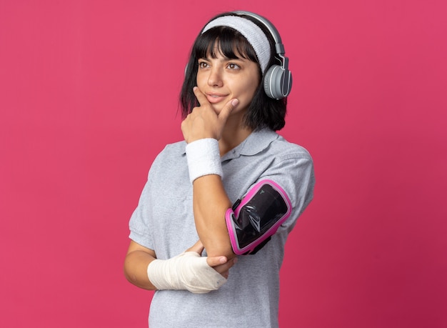 Junges fitness-mädchen mit stirnband mit kopfhörern und armband für smartphone, das mit nachdenklichem gesichtsausdruck beiseite schaut