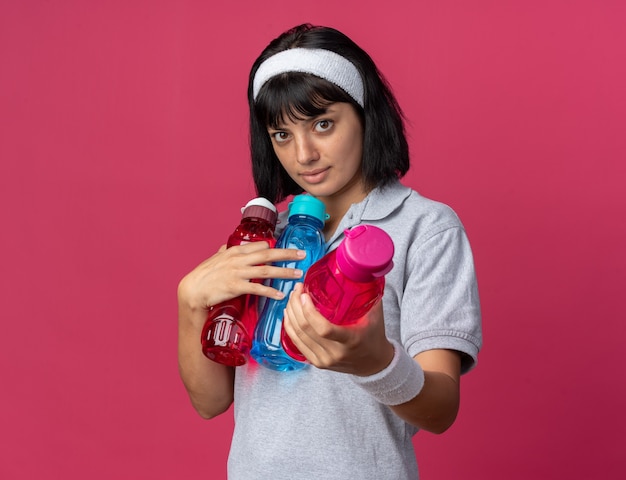 Junges Fitness-Mädchen mit Stirnband mit Wasserflaschen, das einen von ihnen mit ernstem Gesicht über Rosa in die Kamera schaut