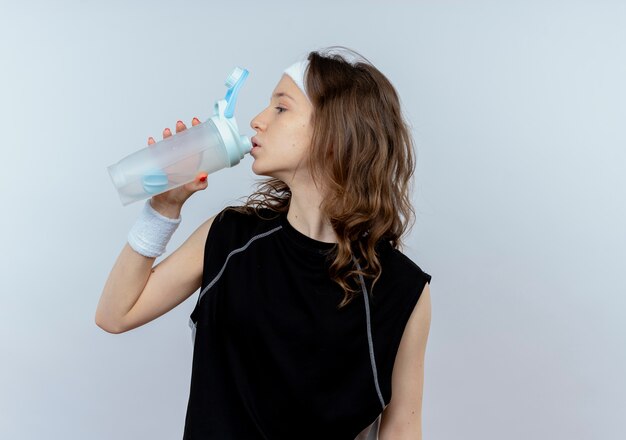 Junges Fitnessmädchen im schwarzen Sportbekleidungs-Trinkwasser nach dem Training, das über weißer Wand steht