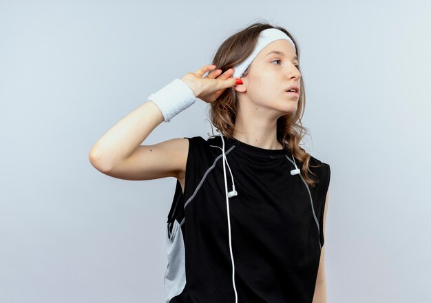 Junges Fitnessmädchen in der schwarzen Sportbekleidung mit dem Stirnband, das beiseite schaut, das ihr Ohr berührt, das versucht, über weißer Wand stehend zu hören