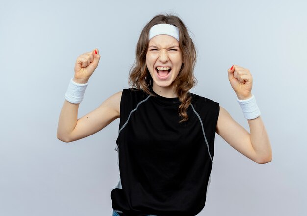 Junges Fitnessmädchen in der schwarzen Sportbekleidung mit dem Stirnband, das die Fäuste glücklich und aufgeregt über weißer Wand zusammenhält