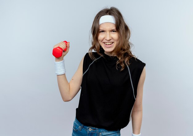 Junges Fitnessmädchen in der schwarzen Sportbekleidung mit dem Stirnband, das Hantel in der erhöhten Hand hält, die sicher steht, über weißer Wand steht