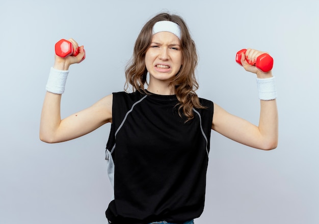 Junges Fitnessmädchen in der schwarzen Sportbekleidung mit dem Stirnband, das mit den Hanteln arbeitet, die angespannt über weißer Wand stehen