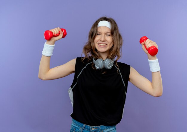 Junges Fitnessmädchen in der schwarzen Sportbekleidung mit dem Stirnband, das mit den Hanteln arbeitet, die über die blaue Wand gespannt stehen