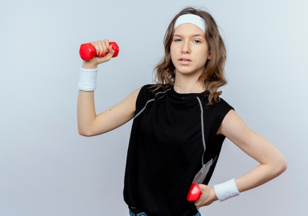 Junges Fitnessmädchen in der schwarzen Sportbekleidung mit dem Stirnband, das mit den Hanteln arbeitet, die zuversichtlich stehen, über weißer Wand stehen