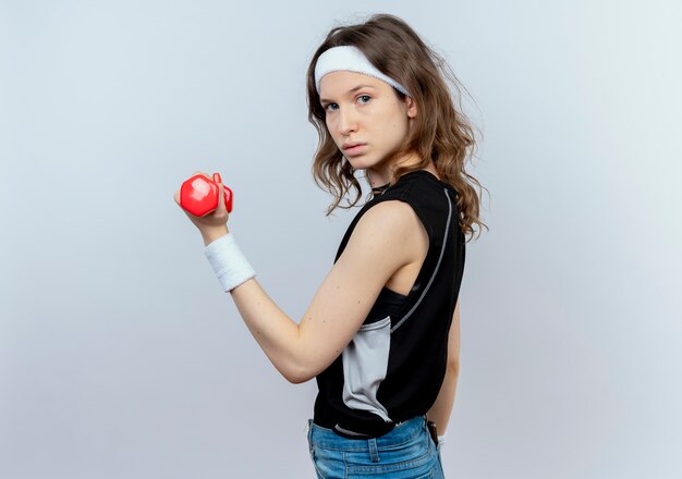 Junges Fitnessmädchen in der schwarzen Sportbekleidung mit dem Stirnband, das mit der Hantel arbeitet, die zuversichtlich steht, über weißer Wand zu stehen