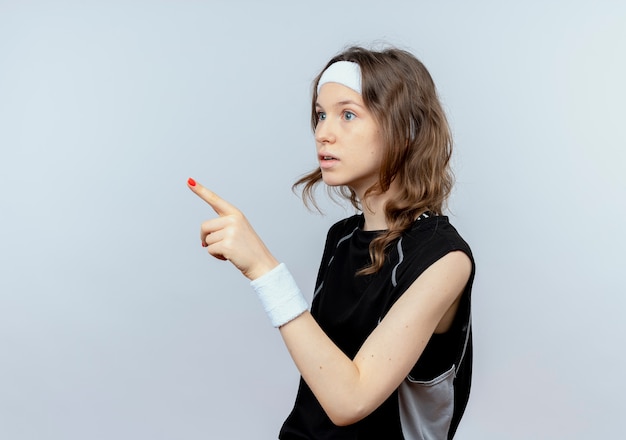 Junges Fitnessmädchen in der schwarzen Sportbekleidung mit dem Stirnband, das verwirrt verwirrt mit dem Finger auf etwas steht, das über weißer Wand steht