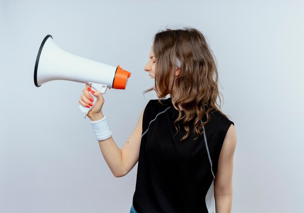 Junges Fitnessmädchen in der schwarzen Sportbekleidung mit dem Stirnband, das zum Megaphon schreit, das über weißer Wand steht