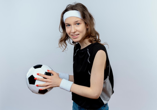 Junges Fitnessmädchen in der schwarzen Sportbekleidung mit dem Stirnband, der Fußball lächelnd steht, der über weißer Wand steht