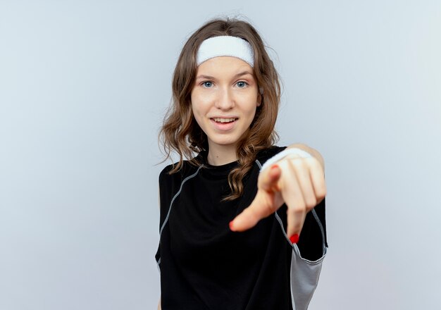 Junges Fitnessmädchen in der schwarzen Sportbekleidung mit dem Stirnband, der mit dem lächelnden Finger zeigt, der über weißer Wand steht