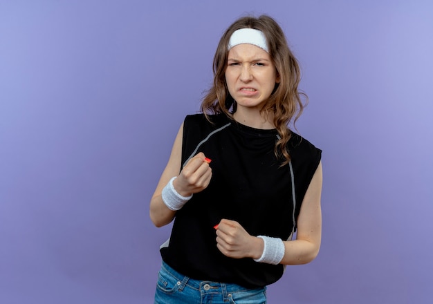 Junges fitnessmädchen in der schwarzen sportbekleidung mit dem stirnband mit den geballten fäusten des verärgerten gesichts, die über der blauen wand stehen