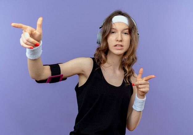 Junges Fitnessmädchen in der schwarzen Sportbekleidung mit dem Stirnband und dem Smartphone-Armband, die mit den Fingern zeigen, die zuversichtlich stehen, über der blauen Wand zu stehen