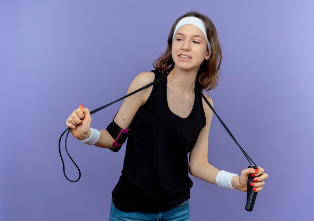 Junges Fitnessmädchen in der schwarzen Sportbekleidung mit Stirnband, das Springseil-Lookign zuversichtlich über der blauen Wand hält