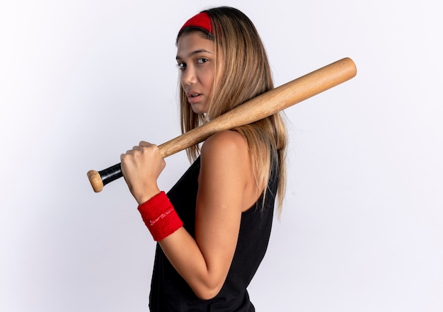Kostenloses Foto junges fitnessmädchen in der schwarzen sportbekleidung und im roten stirnband, die baseballschläger halten, der zuversichtlich steht, über weißer wand steht