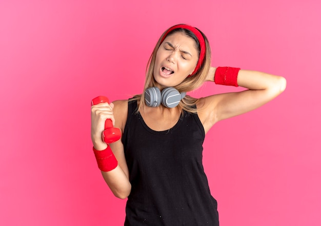 Junges fitnessmädchen in der schwarzen sportbekleidung und im roten stirnband, die hantel halten, die über rosa unzufrieden und frustriert aussieht