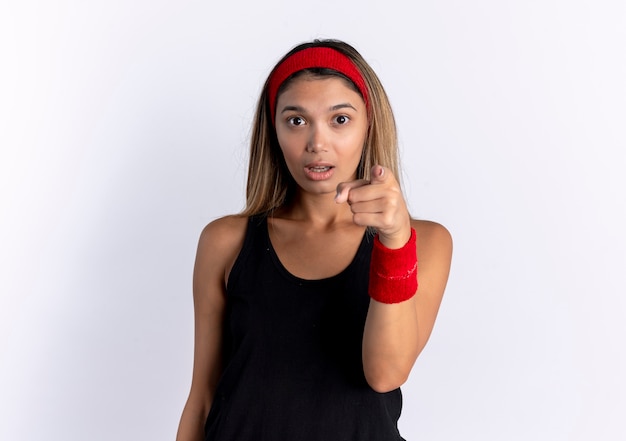 Junges Fitnessmädchen in der schwarzen Sportbekleidung und im roten Stirnband, die mit Zeigefinger zeigen, überrascht, über weißer Wand stehend