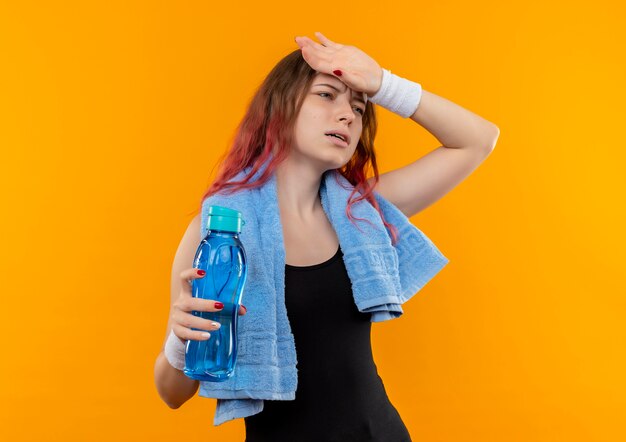 Junges Fitnessmädchen in der Sportbekleidung mit Handtuch auf ihrem Hals, das Flasche Wasser hält, das unwohl schaut, das ihren Kopf berührt über orange Wand steht