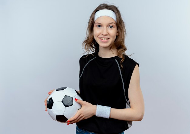 Junges Fitnessmädchen in schwarzer Sportbekleidung mit Stirnband, das Fußball mit Lächeln auf Gesicht steht, das über weißer Wand steht
