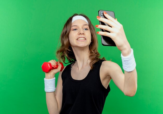 Junges Fitnessmädchen in schwarzer Sportbekleidung mit Stirnband, das Hantel in der Hand hält, macht selfie unter Verwendung des Smartphones, das über grüner Wand steht