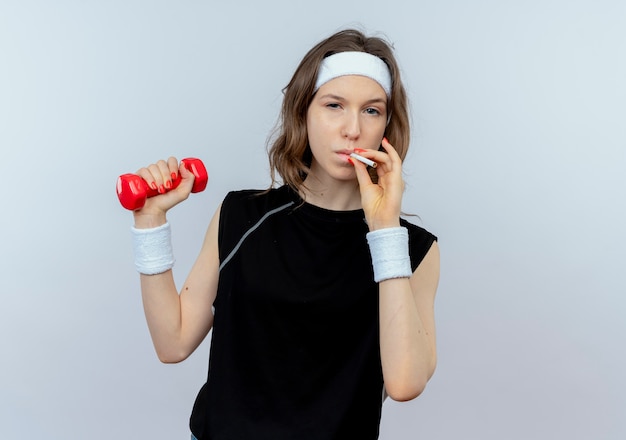 Junges Fitnessmädchen in schwarzer Sportbekleidung mit Stirnband, das mit Hantel und rauchender Zigarette schlechtes Gewohnheitskonzept arbeitet, das über weißer Wand steht