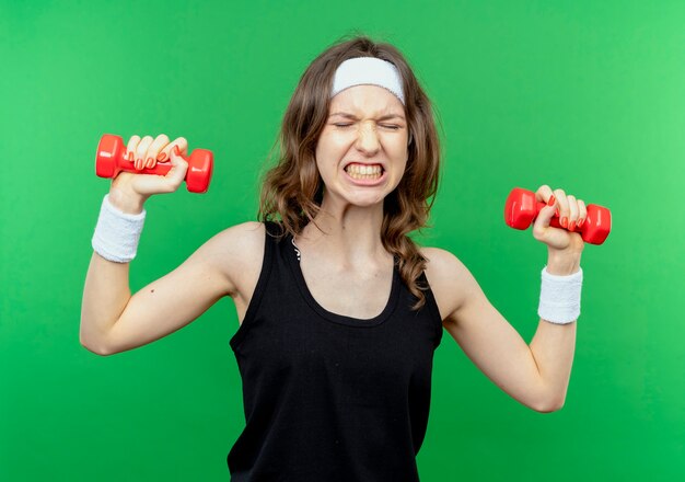 Junges Fitnessmädchen in schwarzer Sportbekleidung mit Stirnband, das mit Hanteln arbeitet, die über Grün angespannt schauen