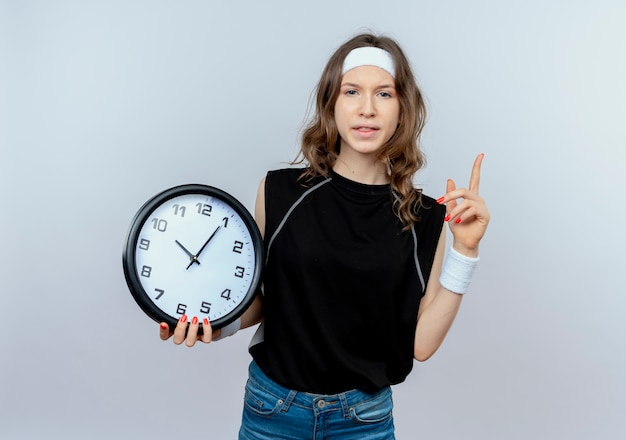 Junges Fitnessmädchen in schwarzer Sportbekleidung mit Stirnband, das Wanduhr hält Zeigefingerwarnung, die über weißer Wand steht