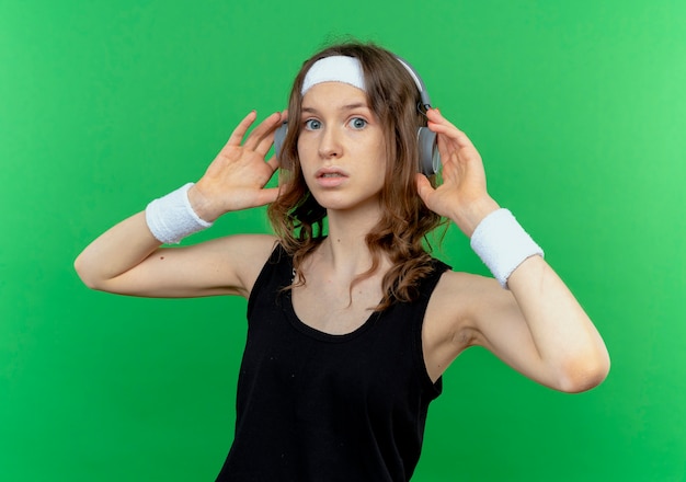 Kostenloses Foto junges fitnessmädchen in schwarzer sportbekleidung mit stirnband mit kopfhörern lookign verwirrt über grüner wand stehend