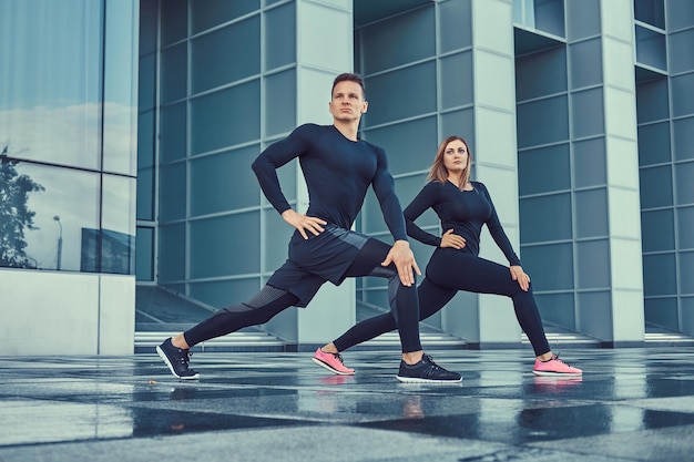 Junges Fitnesspaar in Sportkleidung, das in der modernen Stadt gegen einen Wolkenkratzer Ausfallschritte macht. Gesundes Lebensstilkonzept.