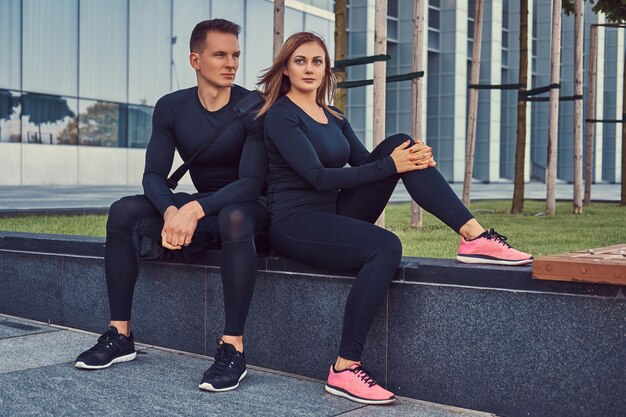 Junges Fitnesspaar in Sportkleidung, sexy blondes Mädchen und ein gutaussehender muskulöser Typ, der auf einer Bank vor einem Wolkenkratzer sitzt.