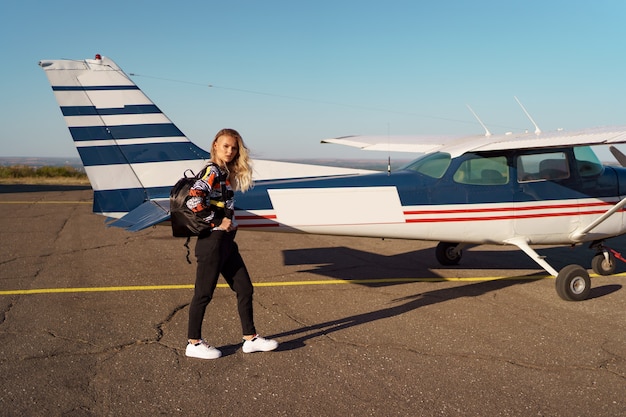 Junges Frauenmodell mit modernem Haarschnitt, das in der Nähe eines Privatflugzeugs posiert und ein trendiges, lässiges Outfit und einen schwarzen Rucksack trägt