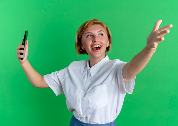 Junges freudiges blondes russisches Mädchen hält Telefon und zeigt vorwärts mit Hand lokalisiert auf grünem Hintergrund mit Kopienraum