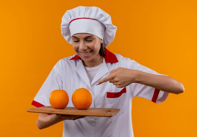 Junges freudiges kaukasisches kochmädchen in der kochuniform hält und zeigt auf orangen auf schneidebrett lokalisiert auf orange raum mit kopienraum