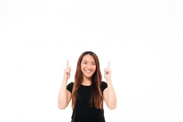 Junges fröhliches asiatisches Mädchen im schwarzen T-Shirt, das mit zwei Fingern nach oben zeigt und Kamera betrachtet