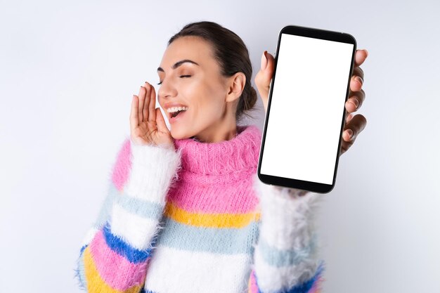 Junges fröhliches Mädchen in einem bunten Pullover auf weißem Hintergrund hält ein großes Telefon im Fokus mit einem leeren weißen Bildschirm