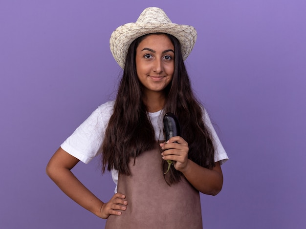 Kostenloses Foto junges gärtnermädchen in der schürze und im sommerhut, die aubergine glücklich und positiv lächelnd stehen über lila wand halten