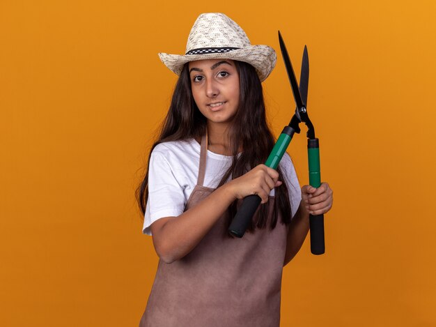 Junges Gärtnermädchen in der Schürze und im Sommerhut, die Heckenscheren halten, die zuversichtlich über orange Wand stehend lächeln