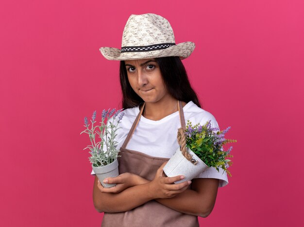 Junges Gärtnermädchen in der Schürze und im Sommerhut, die Topfpflanzen mit wütendem Gesicht halten, das über rosa Wand steht