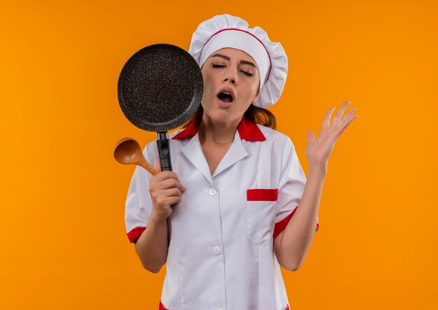 Junges genervtes kaukasisches Kochmädchen in der Kochuniform hält Bratpfanne und Holzlöffel mit geschlossenen Augen lokalisiert auf orange Hintergrund mit Kopienraum