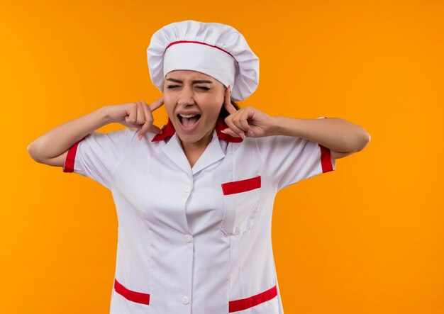 Junges genervtes kaukasisches Kochmädchen in der Kochuniform schließt Ohren mit den Fingern, die auf orange Hintergrund mit Kopienraum isoliert werden