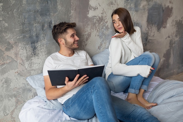 Junges glückliches lächelndes Paar, das auf Bett zu Hause im Lesebuch des lässigen Outfits sitzt, das Jeans, Mann und Frau trägt, die romantische Zeit zusammen verbringen