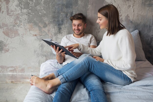 Junges glückliches lächelndes Paar, das auf Bett zu Hause im Lesebuch des lässigen Outfits sitzt, das Jeans, Mann und Frau trägt, die romantische Zeit zusammen verbringen