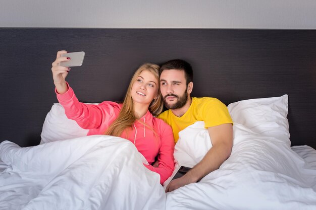 Junges glückliches Paar auf dem Bett, das Selfie mit Telefonkamera macht. Konzept über Technologie und Menschen