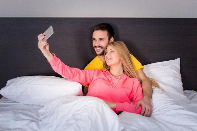 Junges glückliches Paar auf dem Bett, das Selfie mit Telefonkamera macht. Konzept über Technologie und Menschen