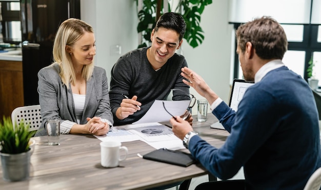 Junges glückliches Paar, das Bedingungen von Hypothekendokumenten liest, während es sich zu Hause mit einem Immobilienmakler trifft. Der Fokus liegt auf dem jungen Mann