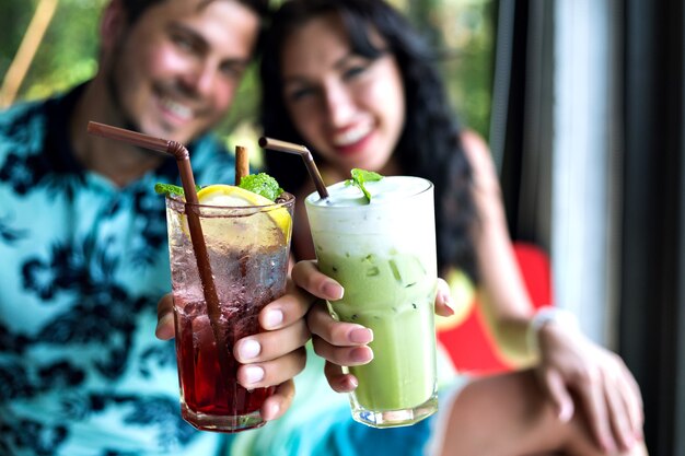 Junges glückliches Paar, das leckere süße Cocktails an der tropischen Bar trinkt, lächelt und Spaß hat, helle Kleidung und positive Gefühle.