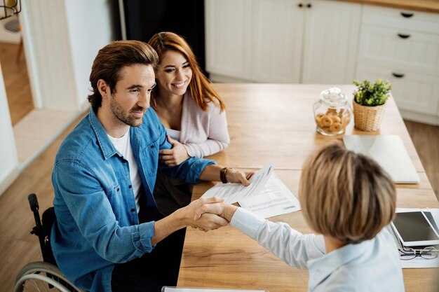 Junges glückliches Paar, das mit seinem Versicherungsagenten eine Vereinbarung über ein Treffen zu Hause trifft. Der Fokus liegt auf dem Mann im Rollstuhl