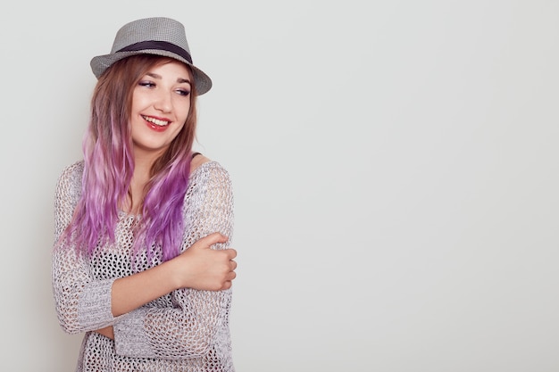 Junges, gut aussehendes Mädchen mit violettem Haar, das Liebe ausdrückt, sich selbst umarmt, mit verträumtem Ausdruck wegschaut, Platz für Werbung kopiert, einzeln auf grauem Hintergrund.