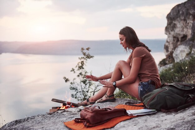 Junges Hipster-Mädchen mit Rucksack, der Sonnenuntergang auf Spitze des Felsenberges genießt.