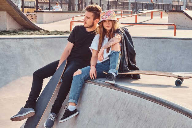 Junges Hipster-Paar, trendig gekleidet, entspannt zusammen mit Skateboards auf einem Skatepark an einem sonnigen Tag.