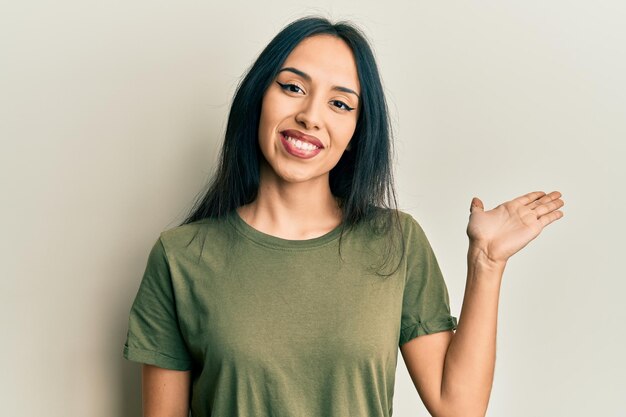 Junges hispanisches Mädchen, das ein lässiges T-Shirt trägt, lächelt fröhlich und zeigt mit der Handfläche in die Kamera.